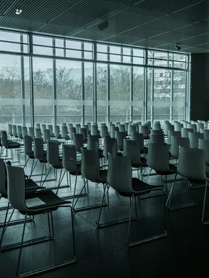Reihen von leeren, modernen Stühlen stehen in einem Konferenz- oder Veranstaltungsraum vor einer großen Fensterfront mit Jalousien. Der Saal ist für eine Tagung oder Präsentation vorbereitet.