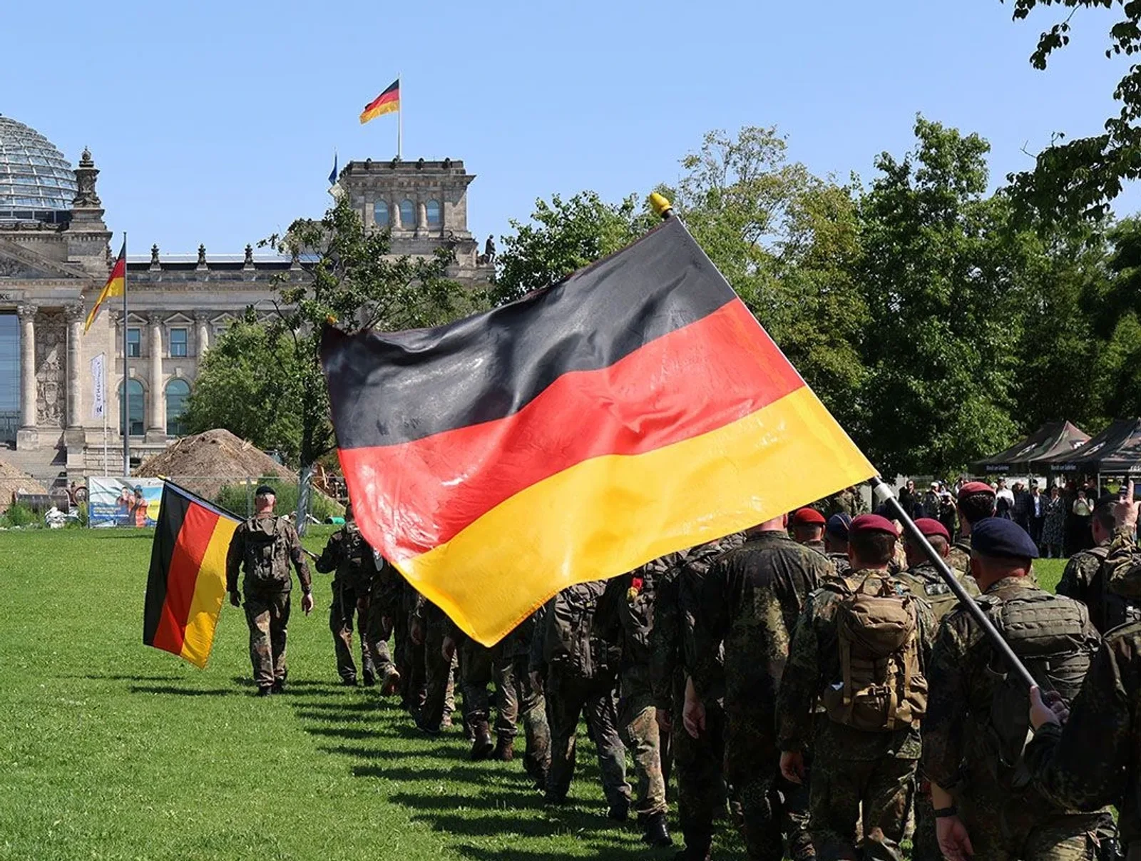 Soldaten der Bundeswehr marschieren in Flecktarn-Uniformen und mit deutschen Nationalflaggen über eine Rasenfläche vor dem Reichstagsgebäude in Berlin. Die Soldaten tragen Rucksäcke sowie Baretts und bewegen sich in einer Formation vor der Kulisse des Parlamentsgebäudes.