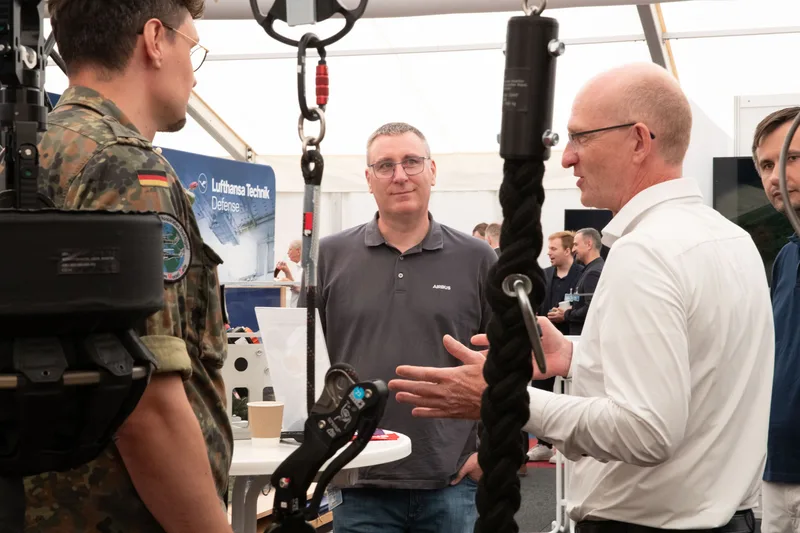 Bundeswehrsoldat im Gespräch mit Airbus-Vertretern vor ausgestellter Abseilausrüstung und einem Stand von Lufthansa Technik Defense