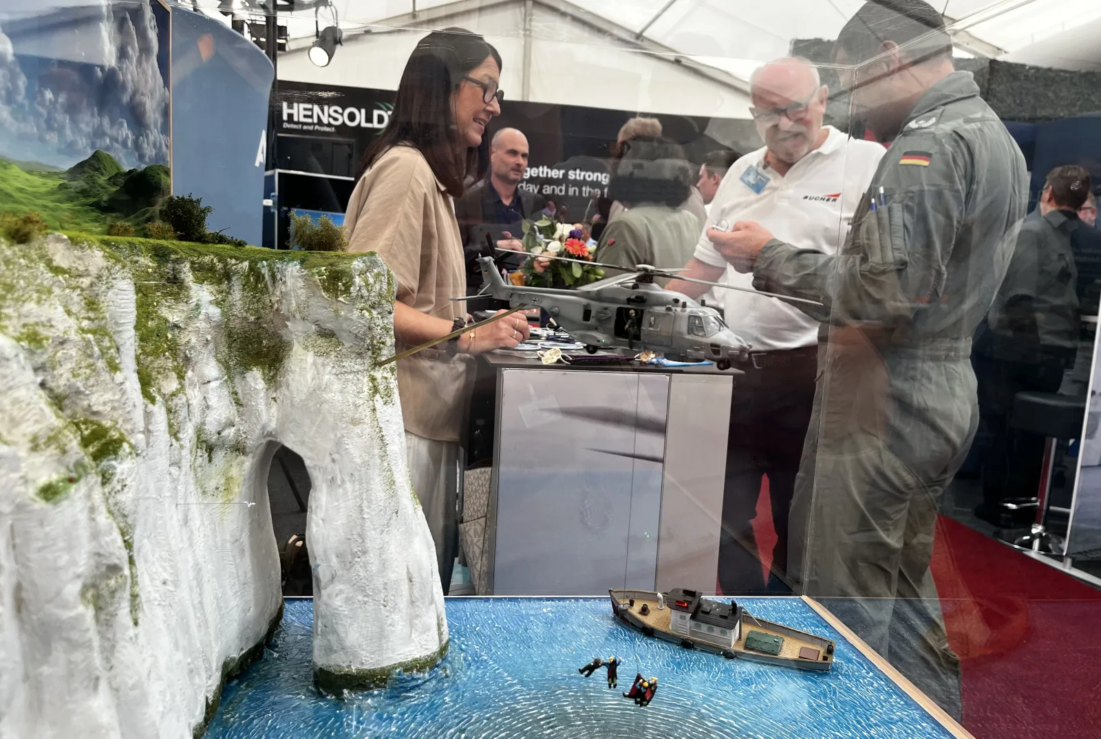 Ein Soldat der Bundeswehr in Fliegerbekleidung und weitere Fachbesucher betrachten auf einer Ausstellung ein Diorama, das ein Modell des Marinehubschraubers NH90 von Airbus Helicopters bei einem maritimen Einsatz zeigt. Im Hintergrund sind Ausstellungsbereiche der Unternehmen Hensoldt und Bucher erkennbar.