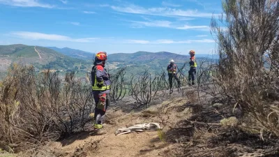 Drei Einsatzkräfte der Feuerwehr in Schutzkleidung begutachten ein von einem Vegetationsbrand betroffenes, hügeliges Gelände. Auf dem staubigen Pfad im Vordergrund liegt ein zusammengerollter Schlauch für die Brandbekämpfung.