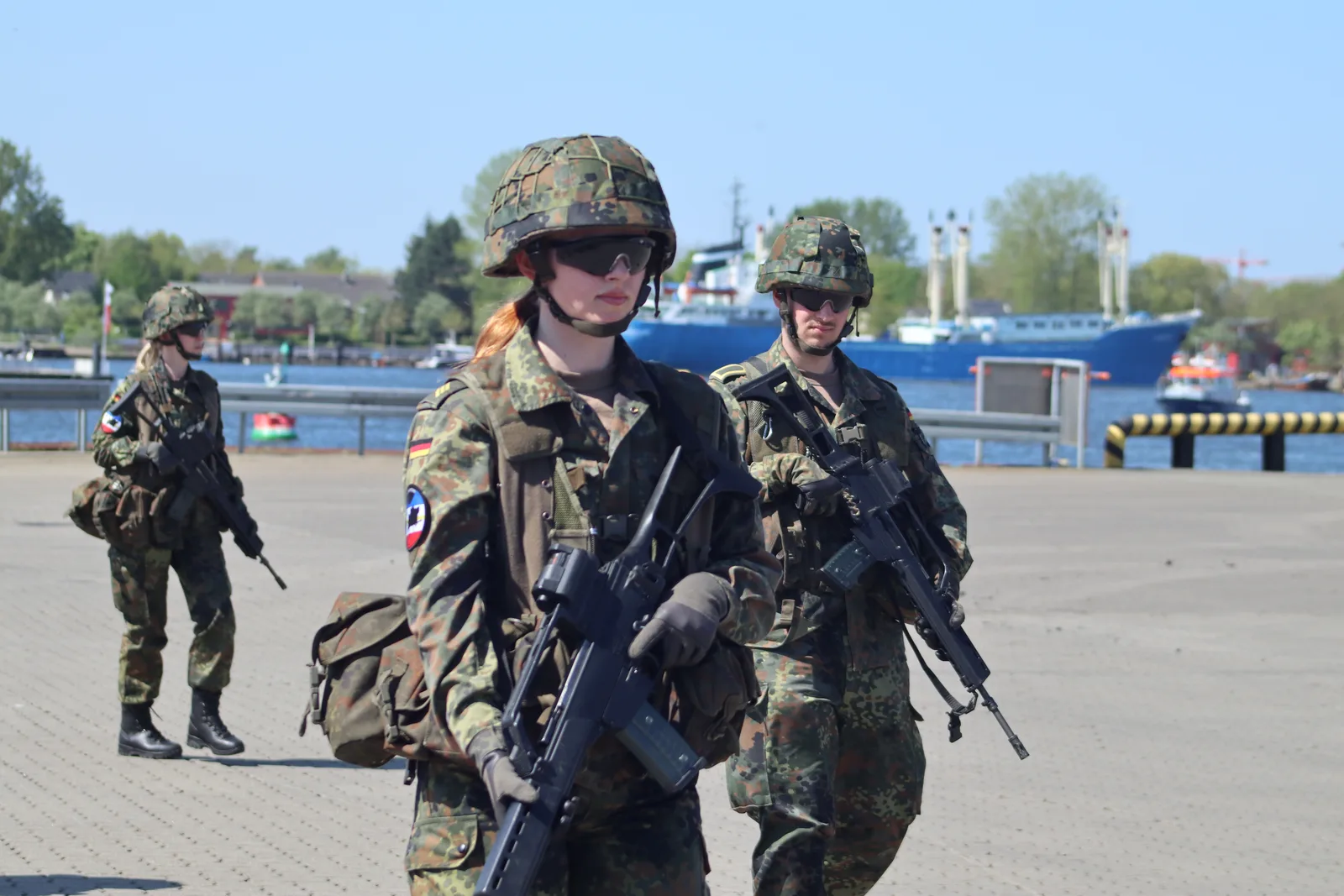 Drei Bundeswehrsoldaten in Flecktarn-Gefechtsbekleidung mit G36-Sturmgewehren bei einer Patrouille an einem Hafenbecken mit Frachtschiff