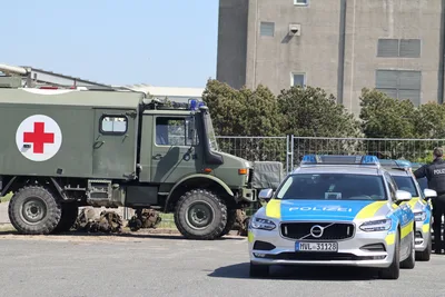 Sanitäts-Unimog der Bundeswehr und Volvo-Streifenwagen der Polizei vor einem Betongebäude mit einem Polizeibeamten rechts