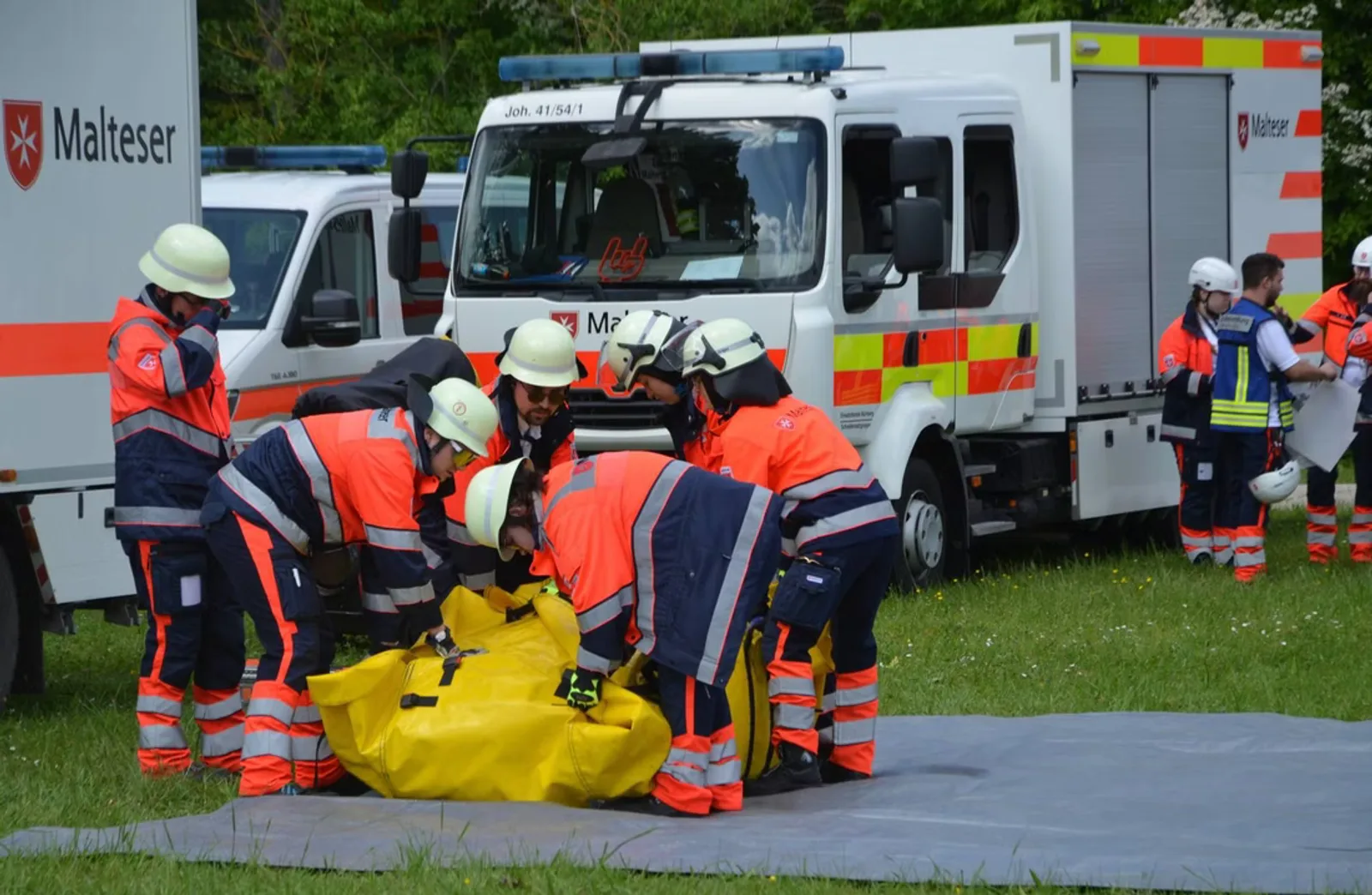 Einsatzkräfte des Malteser Hilfsdienstes bauen während einer Katastrophenschutzübung ein gelbes, aufblasbares Zelt auf einer Wiese auf. Im Hintergrund stehen verschiedene Einsatzfahrzeuge der Hilfsorganisation, darunter ein MAN TGE Transporter.