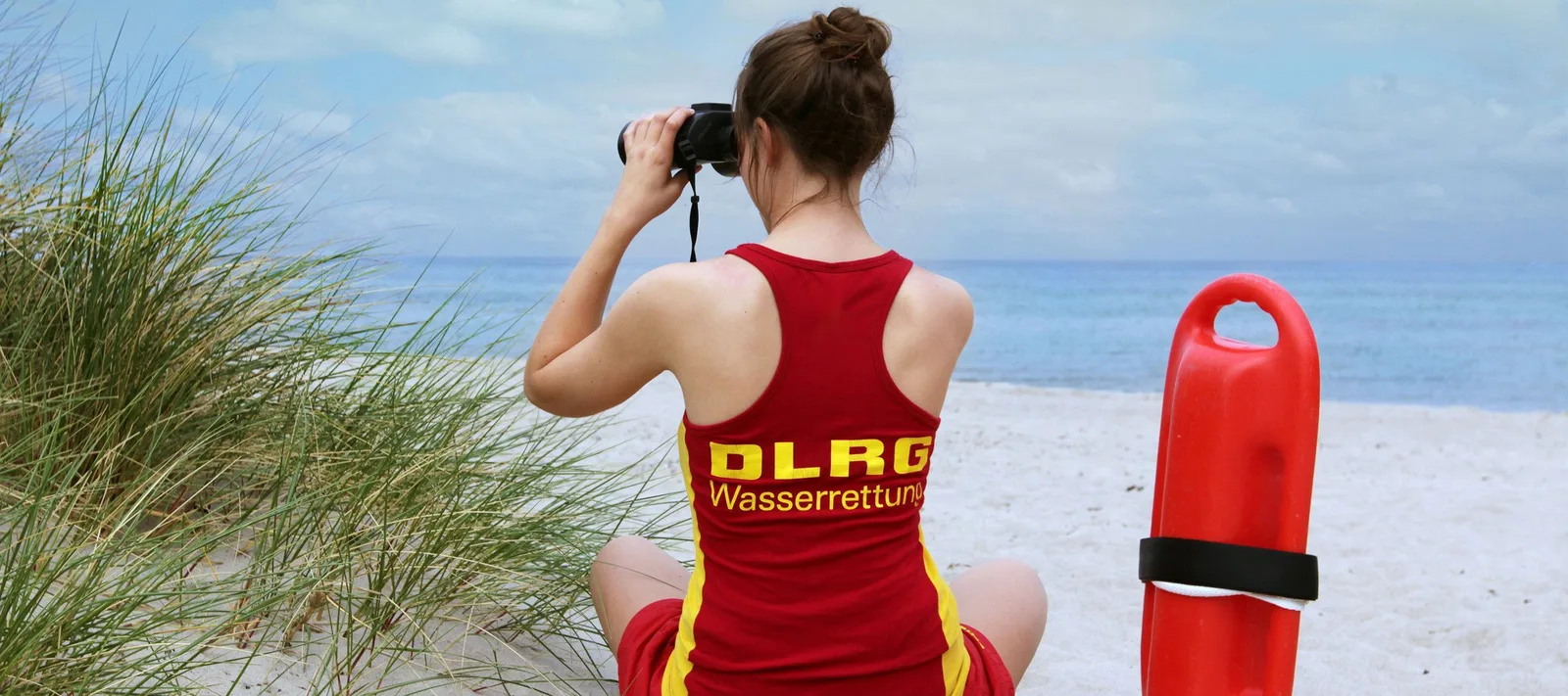 Eine Rettungsschwimmerin der Deutschen Lebens-Rettungs-Gesellschaft (DLRG) beobachtet mit einem Fernglas von einem Sandstrand aus das Meer. Neben ihr im Sand befindet sich eine rote Rettungsboje zur Wasserrettung.