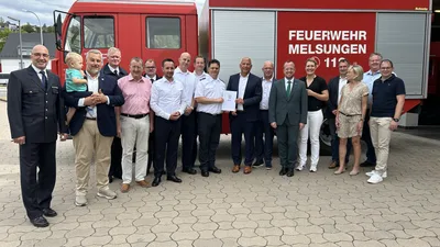 Vertreter aus Politik und Feuerwehr versammeln sich vor einem Einsatzfahrzeug der Freiwilligen Feuerwehr Melsungen zur Übergabe eines offiziellen Bescheids des Landes Hessen. Im Hintergrund ist ein Löschfahrzeug mit einem Aufbau des Herstellers Schmitz zu sehen.