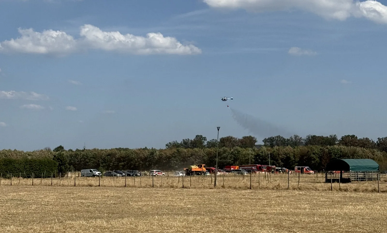Hubschrauber wirft bei einem Waldbrand Wasser aus einem Außenlastbehälter ab, während Einsatzfahrzeuge auf einer Freifläche stehen