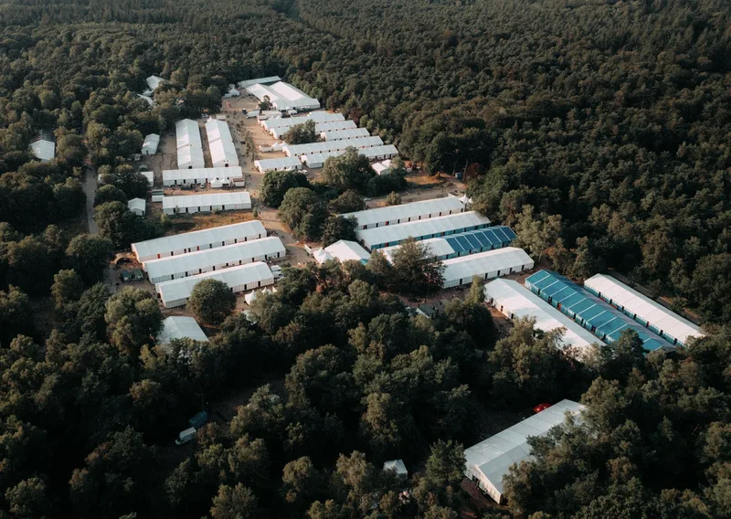 Luftaufnahme einer temporären Zeltstadt mit weißen und blauen Leichtbauhallen in einem Waldgebiet