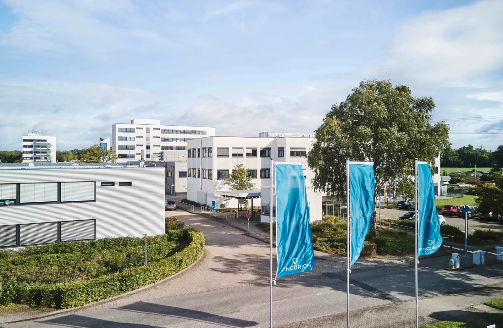 Werksgelände des Technologieunternehmens Vincorion mit Büro- und Betriebsgebäuden sowie drei blauen Flaggen mit Firmenlogo im Vordergrund