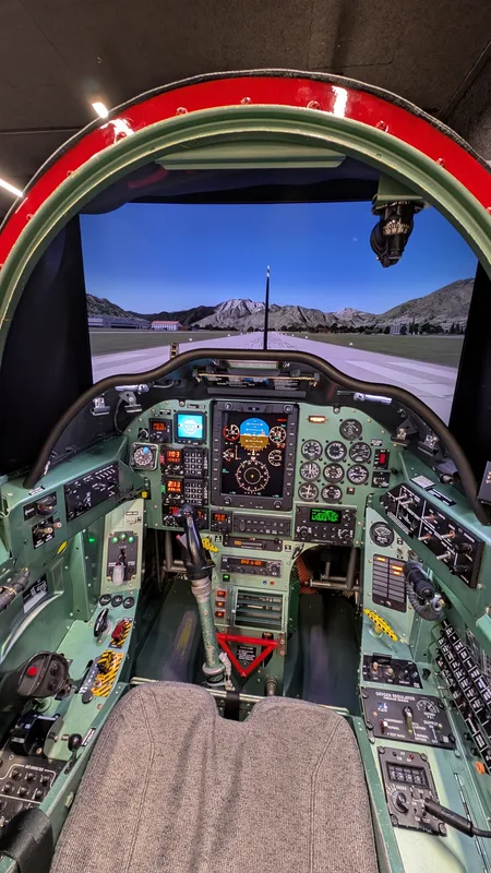 Cockpit eines Flugsimulators der Luftwaffe mit Instrumententafeln, Steuerknüppel und projizierter Landebahn vor Gebirgskulisse