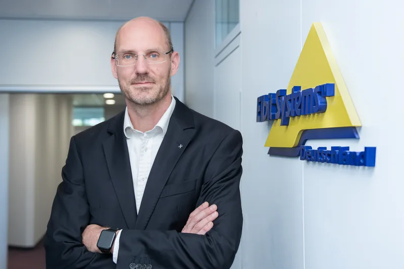 Michael Menges von Elbit Systems Deutschland steht mit verschränkten Armen vor dem Firmenlogo in einem Bürogebäude