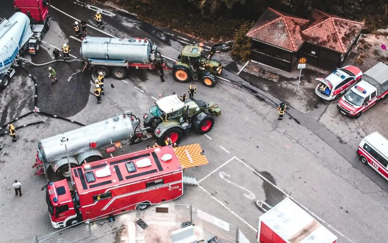 Drohnenaufnahme von Feuerwehrfahrzeugen und Fendt-Traktoren mit Tankanhängern bei einem Löschwassereinsatz auf einer Freifläche