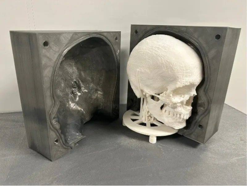 Weißes 3D-gedrucktes Schädelmodell neben einer geöffneten zweiteiligen Gussform für ein vollständiges Kopfmodell