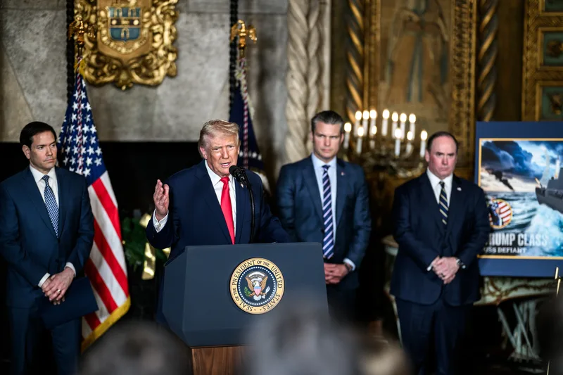 Donald Trump spricht an einem Rednerpult mit US-Präsidentensiegel neben Senator Marco Rubio vor einer US-Flagge und einem Kriegsschiff-Entwurf
