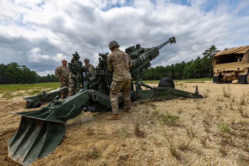 Soldaten der US-Nationalgarde bedienen eine M777-Feldhaubitze auf einem sandigen Übungsplatz mit Zugfahrzeug im Hintergrund