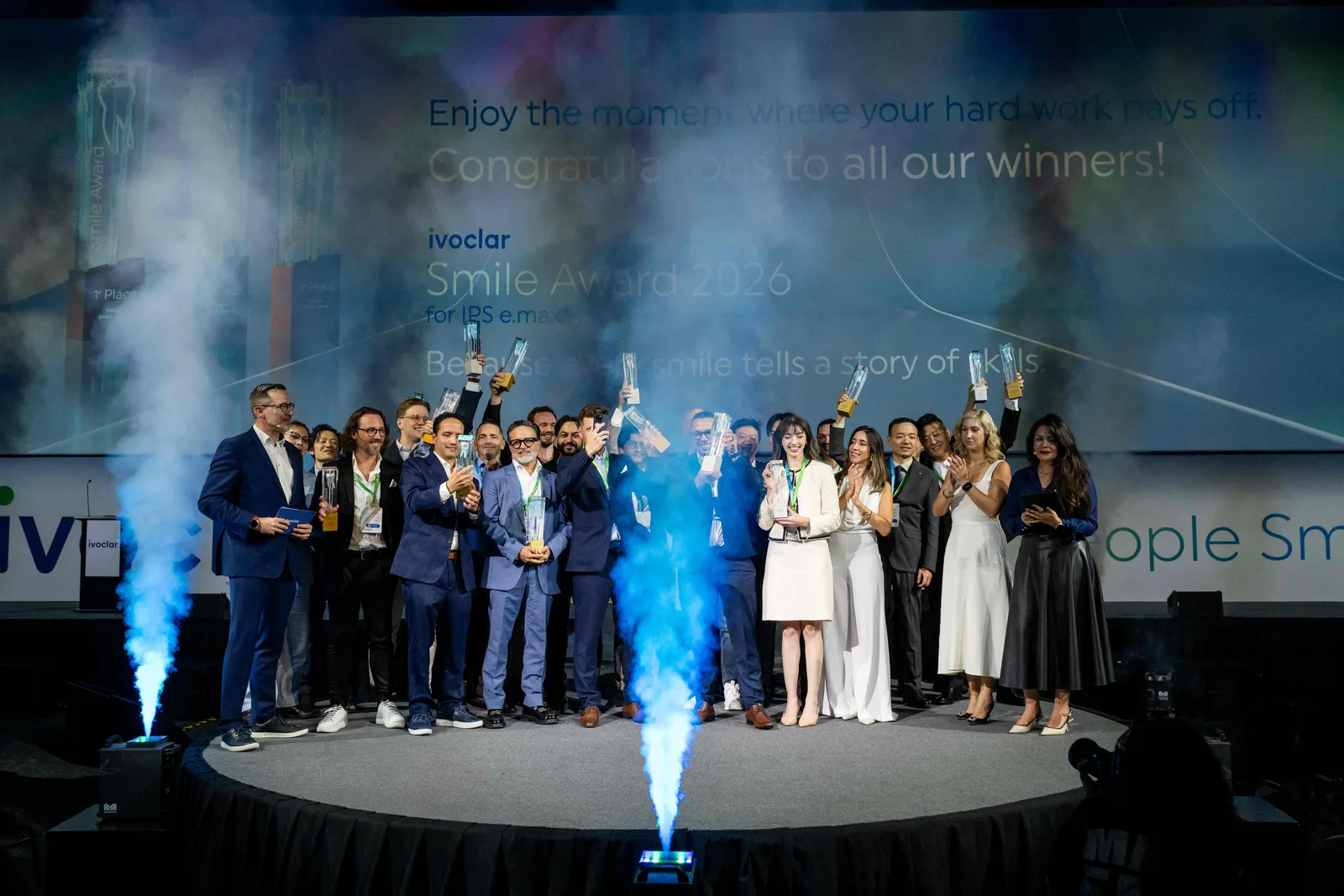 Preisträger posieren mit Trophäen auf der Bühne des Ivoclar Smile Award vor einer Firmenleinwand mit blauen Raucheffekten