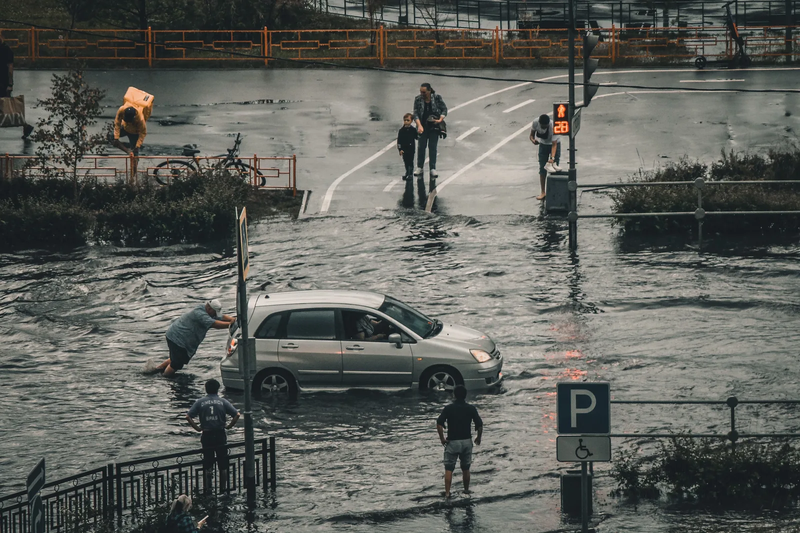 Hochwasser: Ein Mann schiebt einen Nissan Almera Tino durch eine überflutete Straße nach Starkregen, beobachtet von Passanten am Straßenrand.
