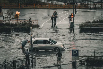 Hochwasser: Ein Mann schiebt einen Nissan Almera Tino durch eine überflutete Straße nach Starkregen, beobachtet von Passanten am Straßenrand.
