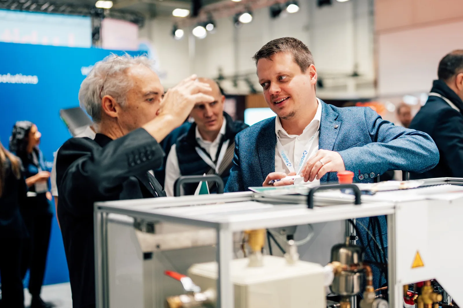 E-world: Zwei Männer im Gespräch an einem technischen Demonstrationsgerät mit Rohrleitungen auf einem Messestand der Firma epilot