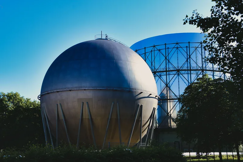 Gas: Kugelgasbehälter und Gasometer mit Stahlgitterkonstruktion auf einem bewaldeten Industriegelände unter blauem Himmel