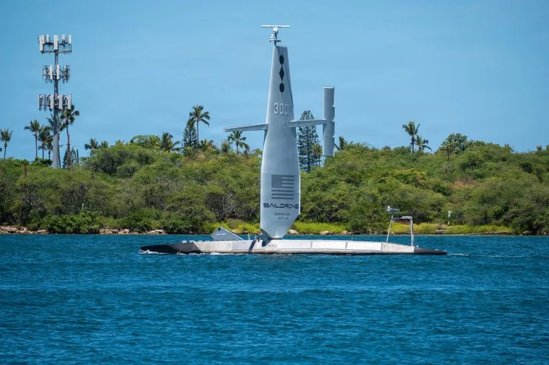 Unbemanntes Oberflächenfahrzeug Saildrone Surveyor mit Starrsegel und Sensoren auf dem Wasser vor einer bewaldeten Küste
