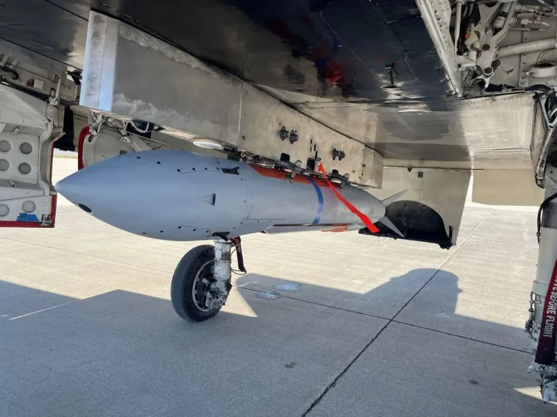 Grauer Sensorbehälter an der Außenlaststation eines Kampfflugzeugs auf einem Flugplatz mit sichtbarem Fahrwerk im Vordergrund