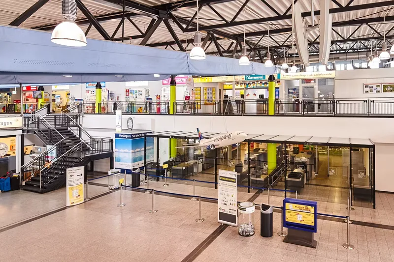 Leere Sicherheitskontrollstelle im Terminal des Flughafens Paderborn-Lippstadt mit Reisebüros und einem Modellflugzeug im Hintergrund