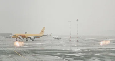 Drohne: Gelbes DHL-Frachtflugzeug mit mobiler Einstiegstreppe auf einem nassen, nebligen Flughafen-Vorfeld