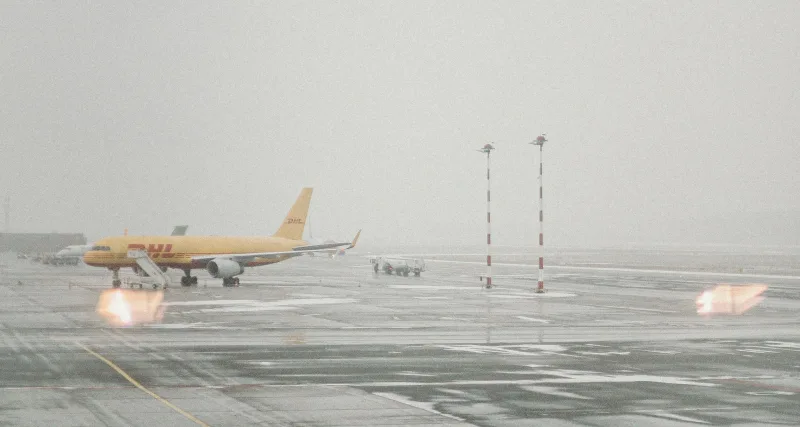 Drohne: Gelbes DHL-Frachtflugzeug mit mobiler Einstiegstreppe auf einem nassen, nebligen Flughafen-Vorfeld