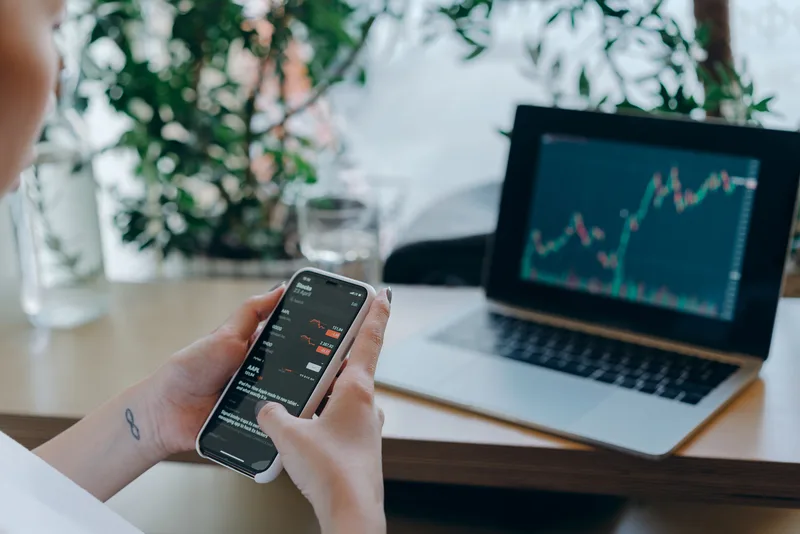 LKA: Person analysiert Finanzdaten und Aktienkurse auf einem Apple iPhone und einem Laptop mit geöffnetem Diagramm vor Zimmerpflanzen
