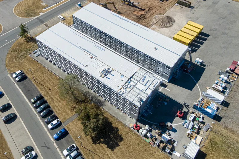 Luftaufnahme zweier dreigeschossiger Containergebäude auf einem Baustellengelände mit DHL-Containern, Fahrzeugen und Baumaschinen
