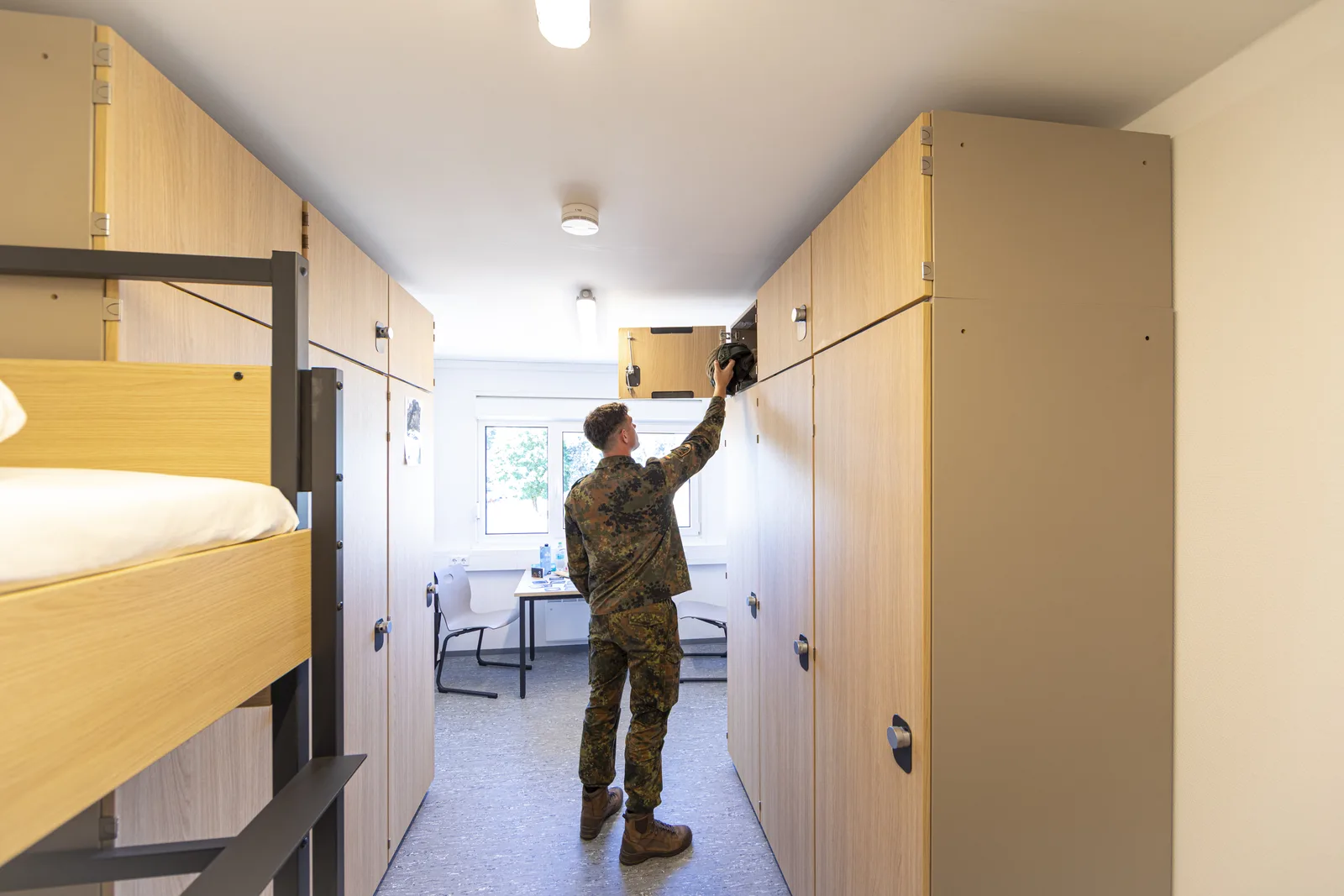Unterkünfte: Bundeswehr-Soldat in Flecktarn-Uniform verstaut Ausrüstung in einem Holzspind in einer modernisierten Stube mit Etagenbetten