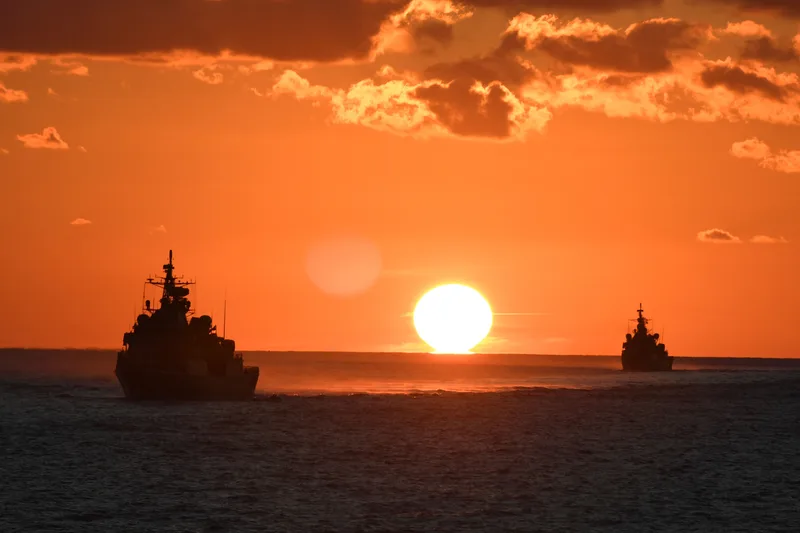 Zwei Kriegsschiffe fahren bei Sonnenuntergang in Formation auf offener See als dunkle Silhouetten vor orangefarbenem Himmel