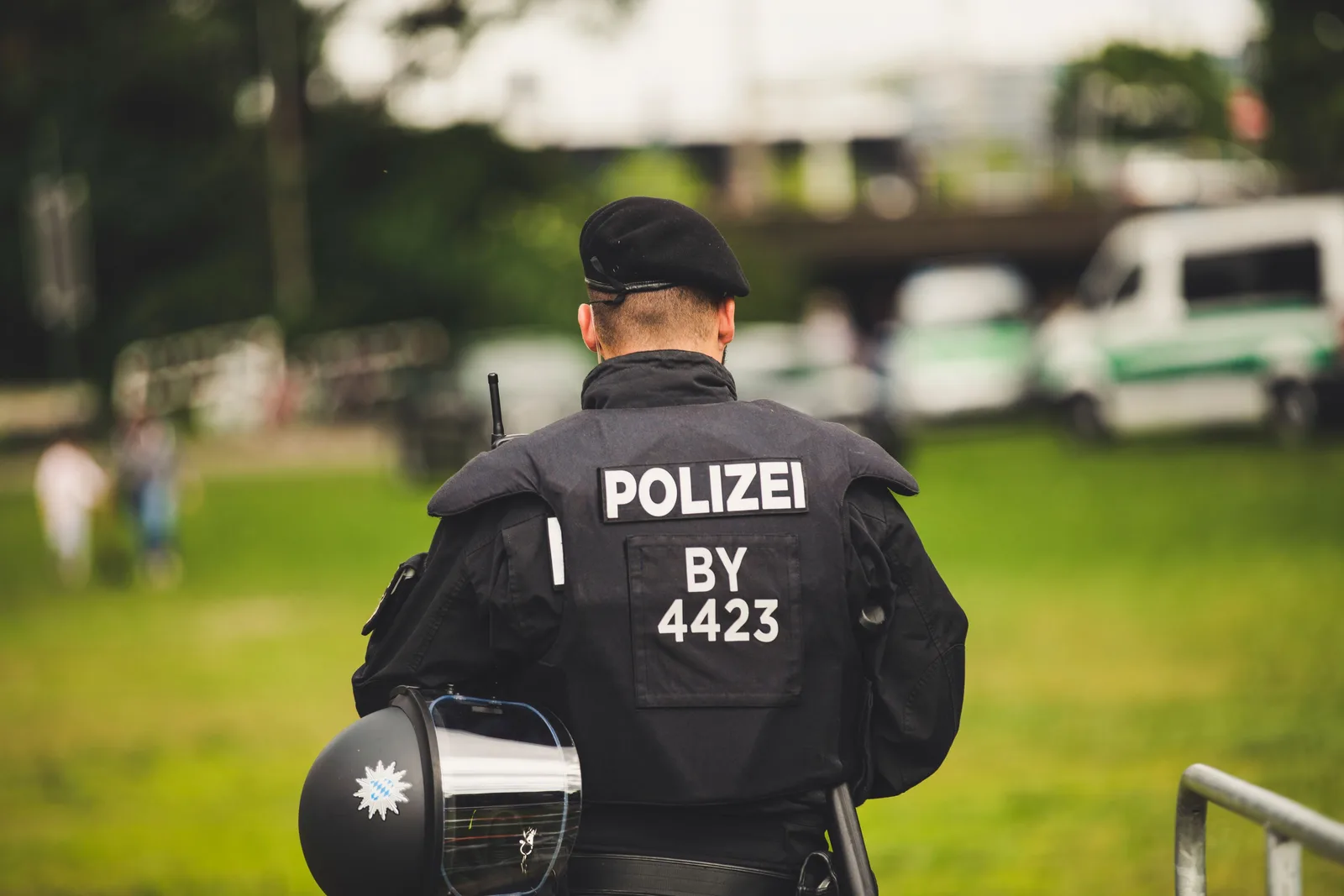 Bayerischer Polizeibeamter in schwarzer Schutzausrüstung mit Barett und Einsatzhelm steht vor parkenden Polizeifahrzeugen