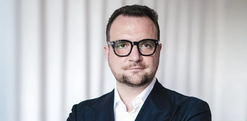 Mann mit Brille, weißem Hemd und dunklem Sakko vor neutralem Hintergrund