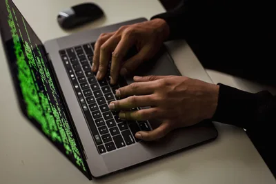 Bayern: Hände tippen auf einem Apple MacBook Pro mit grünem Programmcode auf dem Bildschirm neben einer Computermaus auf hellem Schreibtisch