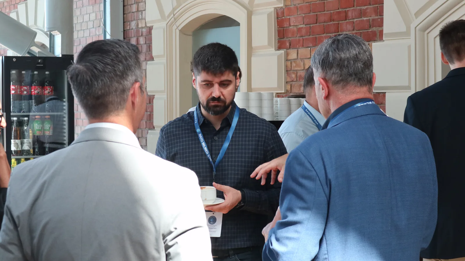 "Golden Hour doesn't work here" – Ukrainian military doctor wakes up NATO – Teilnehmer in Anzügen unterhalten sich bei einer Konferenz des NATO MILMED COE, ein Mann mit blauem Schlüsselband hält eine Kaffeetasse