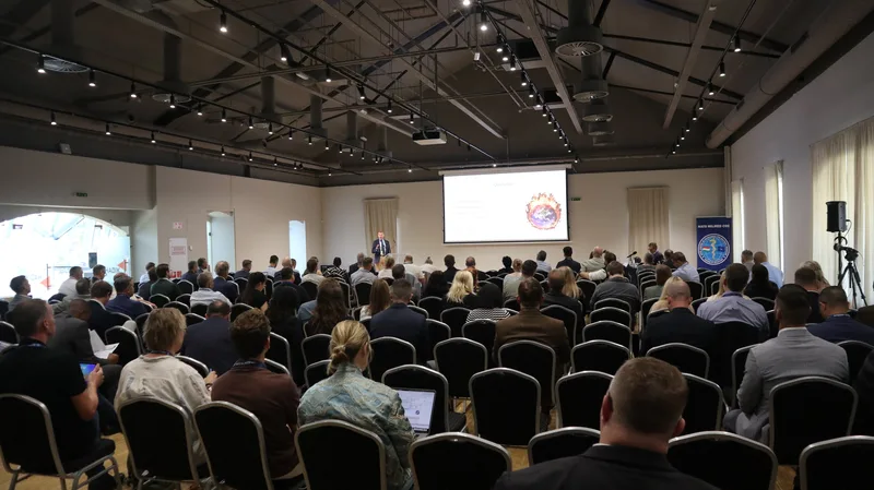 Referent hält Vortrag vor Fachpublikum im Saal mit Projektionsfläche und Rollbanner des NATO MILMED COE