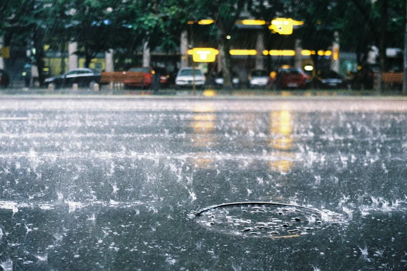 Tool: Starker Regen fällt auf eine nasse Asphaltstraße mit einem runden Kanaldeckel im Vordergrund und parkenden Fahrzeugen im Hintergrund
