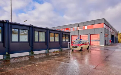 Modulare Rettungswache von Adapteo neben einem festen Feuerwehrgebäude mit geparktem Rettungsfahrzeug auf Basis eines Volkswagen T4