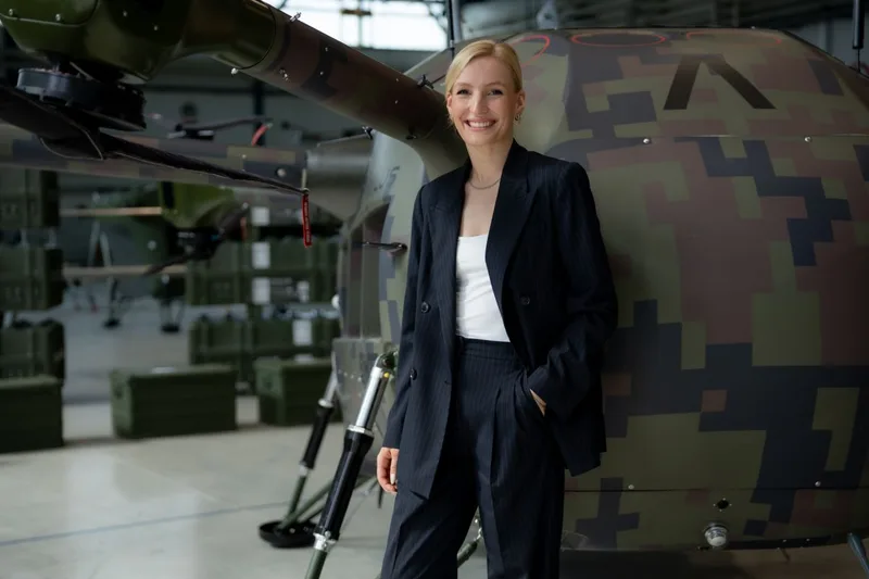 Frau im Hosenanzug steht lächelnd vor einem unbemannten Airbus-Hubschrauber mit digitalem Tarnmuster in einer Lagerhalle