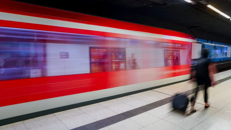 Explosion: Ein rot-weißer Personenzug fährt mit hoher Geschwindigkeit durch einen unterirdischen Bahnhof, während eine Person mit Koffer vorbeiläuft