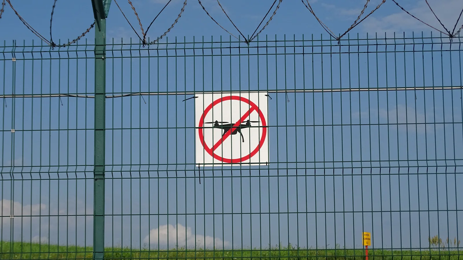 Grüner Sicherheitszaun mit Stacheldraht und einem Hinweisschild für ein Drohnen-Flugverbot an einem abgesperrten Gelände