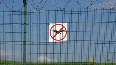 Grüner Sicherheitszaun mit Stacheldraht und einem Hinweisschild für ein Drohnen-Flugverbot an einem abgesperrten Gelände