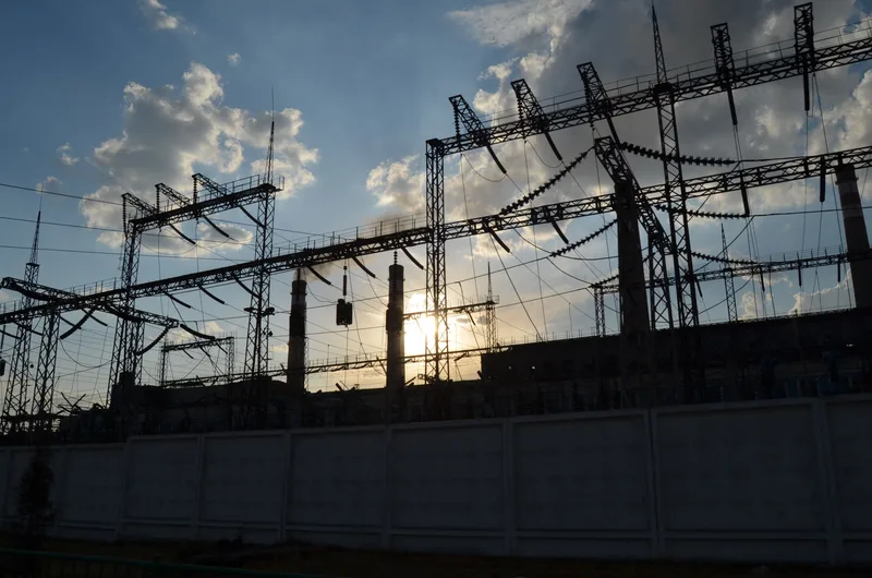 Umspannwerk mit Hochspannungsmasten vor der Silhouette eines Kraftwerks im Gegenlicht der tiefstehenden Sonne