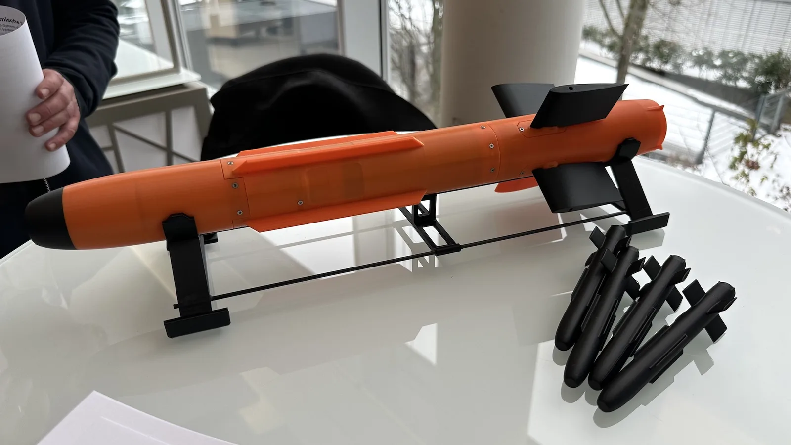 NRW.BANK: Orangefarbenes Lenkflugkörper-Modell auf Ständer und vier schwarze Präzisionsmunitions-Modelle auf einem weißen Tisch