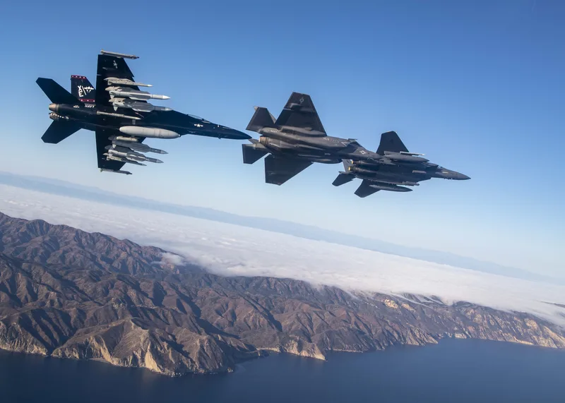 Eine Boeing F/A-18 Super Hornet im Rückenflug in Formation mit zwei Lockheed Martin F-35 Lightning II über einer Küste