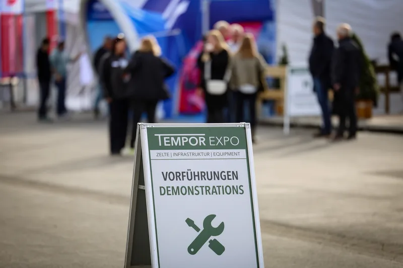 Hinweisschild für Vorführungen auf der Fachmesse Tempor Expo vor unscharfen Besuchern auf dem Ausstellungsgelände