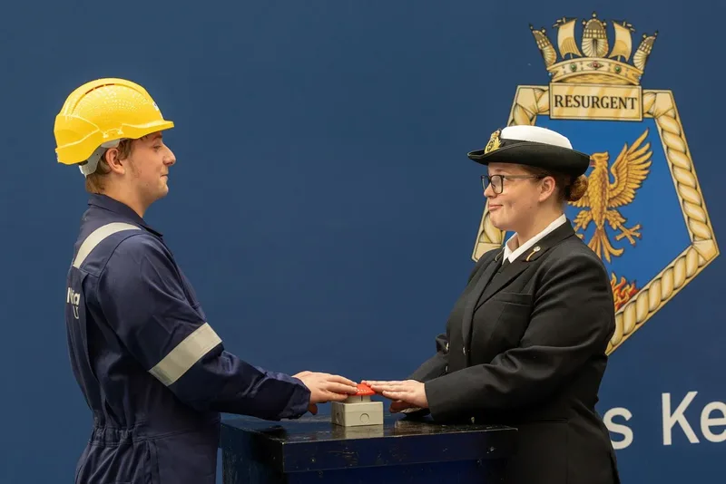 Ein Arbeiter in blauem Overall mit Schutzhelm und eine britische Soldatin betätigen gemeinsam einen roten Druckknopf an einem Podest