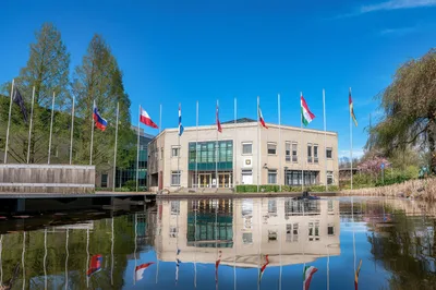CIMC Gebäude des CCOE in Den Haag mit davor wehenden Flaggen der Partnernationen und Spiegelung im Teich unter blauem Himmel